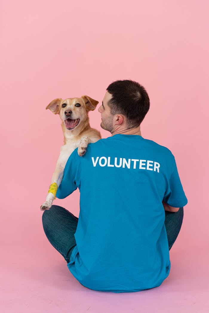 menu-16 A volunteer wearing a blue shirt cuddles a happy dog in a studio with a pink background.
