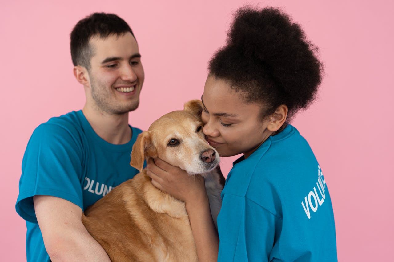 Two volunteers happily hugging a dog against a light pink backdrop.